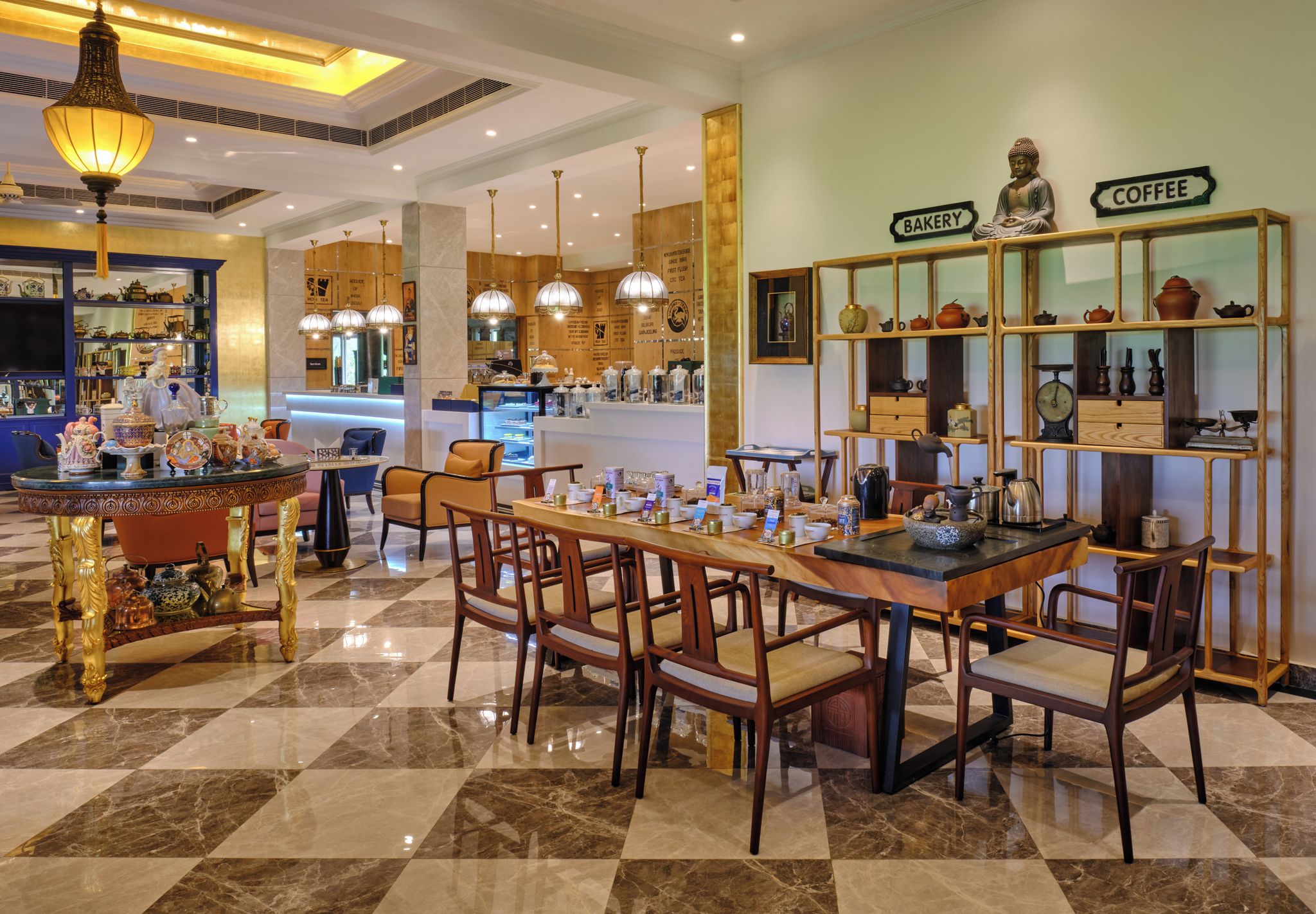 A hotel lobby with a checkered floor, a central dining table with chairs, a coffee station, and a bakery display.