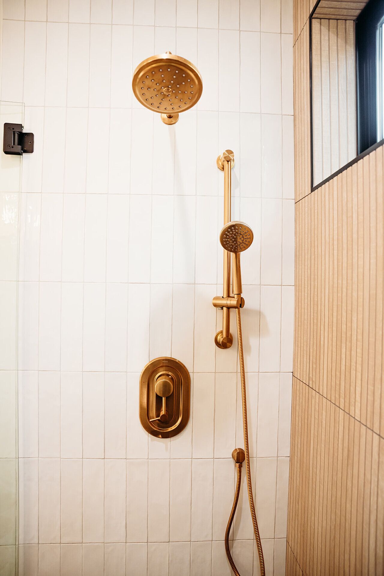 A bathroom with white tiled walls, a gold showerhead, a handheld showerhead, and a gold faucet.