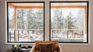 A room with two large windows overlooking a forested area. There is a wicker chair with a brown cushion in the foreground. - Kestrel House At The Running Y (Klamath Falls)