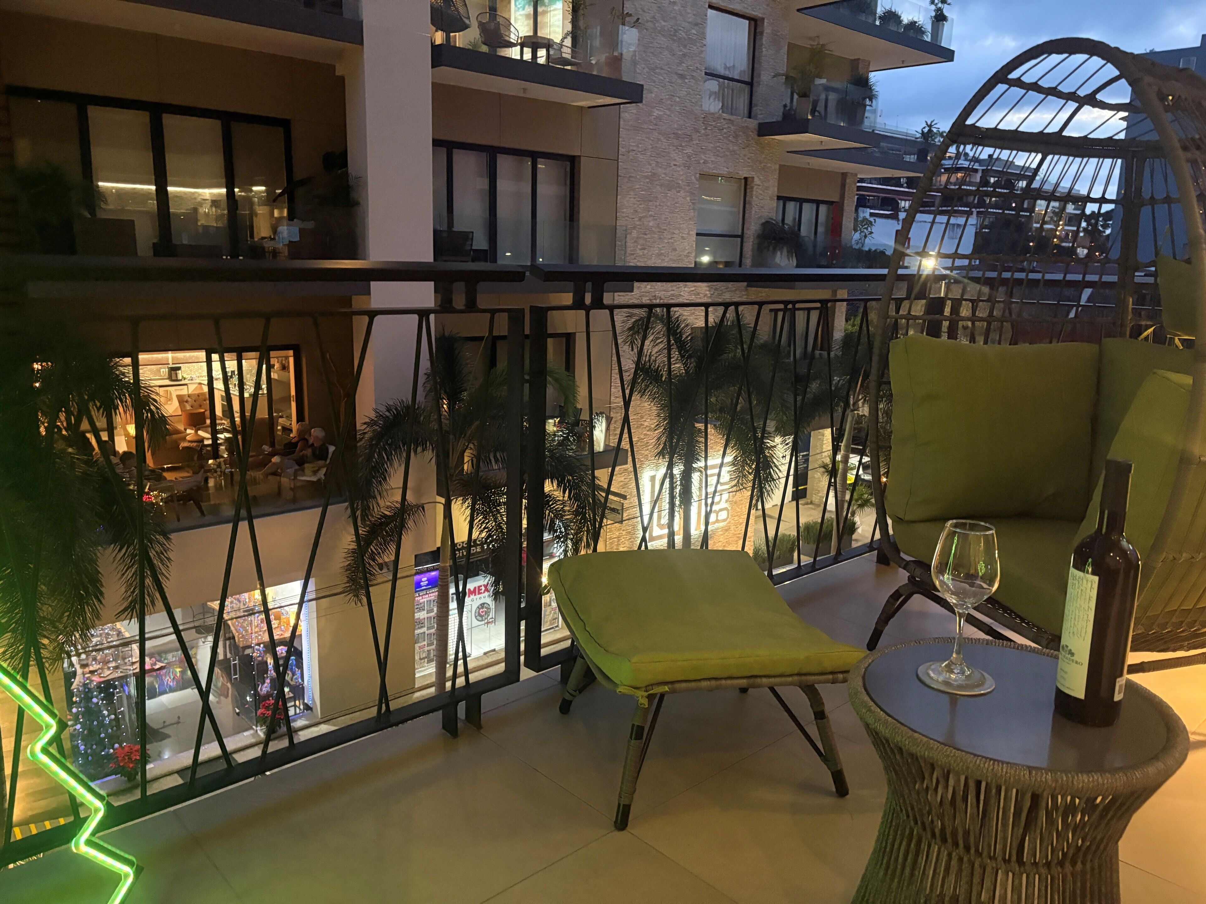 A balcony with a wicker chair, a small table with a wine glass and bottle, and a view of a cityscape with palm trees.