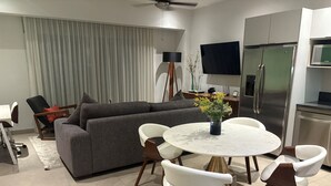 Ein modernes Wohnzimmer mit einem grauen Sofa, einem runden Esstisch mit weißen Stühlen, einem flachbildfernseher an der Wand und einem Edelstahlkühlschrank.
