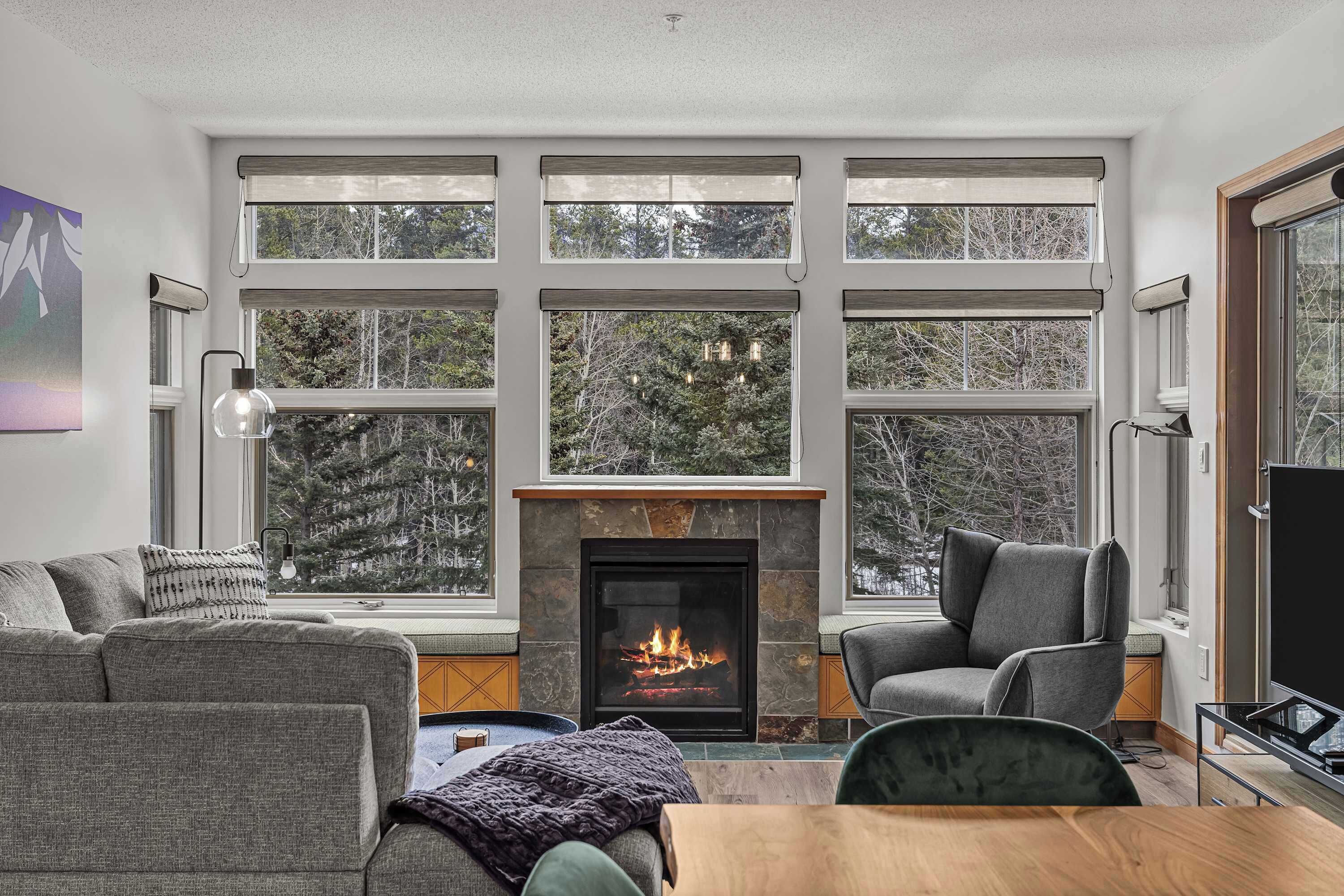 A cozy living room with a fireplace, large windows, a sofa, a chair, and a TV.