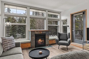 A modern living room with a fireplace, large windows, and a cozy seating area. - Tranquil Condo In The Trees, Spacious 2Bd Hot Tub & Pool (Canmore)
