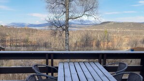 En träveranda med två fåtöljer av rotting och ett bord, med utsikt över ett skogslandskap och berg i fjärran.