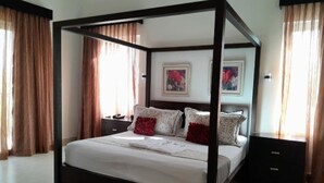 Un lit à baldaquin avec une structure en bois, une literie blanche et des coussins décoratifs. Il y a deux tables de chevet avec des lampes, un téléviseur fixé au mur et des œuvres d’art au-dessus du lit.