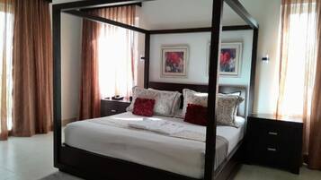 A four-poster bed with a wooden frame, white bedding, and decorative pillows. There are two nightstands with lamps, a wall-mounted TV, and artwork above the bed.