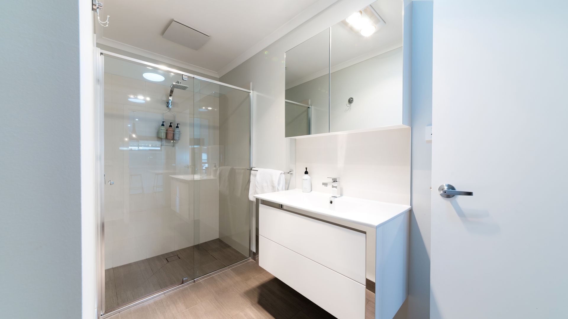 Un baño moderno con cabina de ducha de vidrio, un tocador blanco con espejo y una puerta a la derecha.