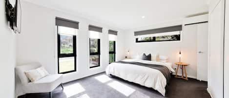 Un dormitorio moderno con una cama, una silla, una mesita de noche y ventanas amplias.
