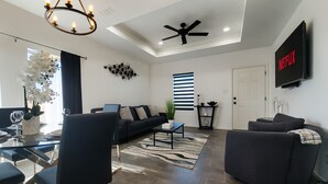 Una sala de estar moderna con un sofá, sillones, una mesa de centro y un comedor con una mesa de vidrio y sillas. Hay un ventilador de techo, una araña y un televisor montado en la pared.