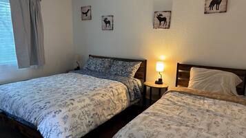 A bedroom with two beds, a window with curtains, a bedside table with a lamp, and wall art depicting animals.