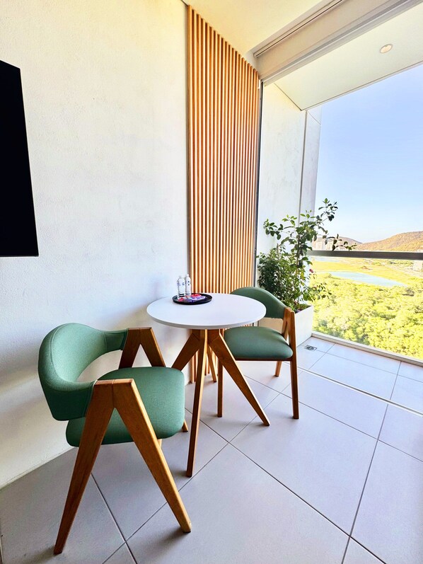 Een moderne kamer met een ronde tafel, twee groene stoelen, een houten vloer, een witte muur en uitzicht op een landschap door een raam.