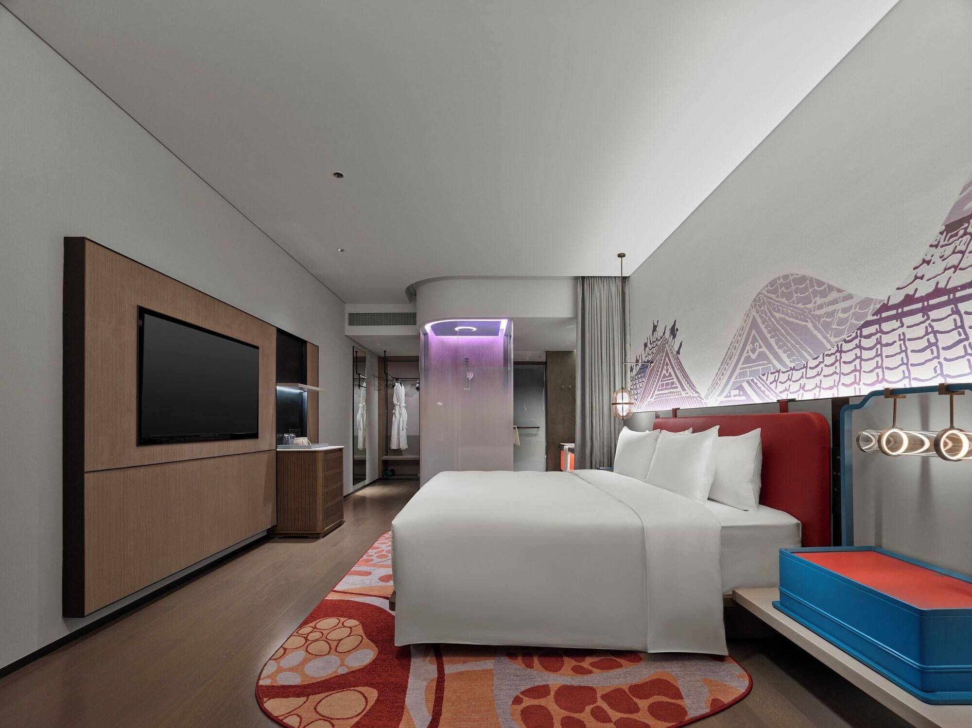A modern hotel room with a large bed, a flat-screen TV, a vanity with a mirror, and a decorative wall mural.