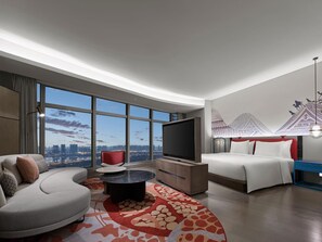 A modern hotel room with a curved sofa, a round coffee table, a flat-screen TV, a bed with white bedding, and a cityscape view through large windows. - Dhawa Xi'an Chanba (Xi'an)