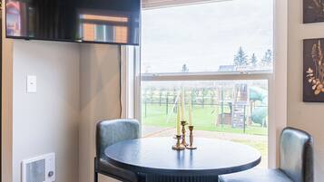 A dining area with a round table, two chairs, a TV mounted on the wall, and a window with a view of a playground.