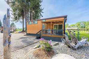 A cabin with a deck, surrounded by gravel and rocks, with trees and a grassy area in the background.