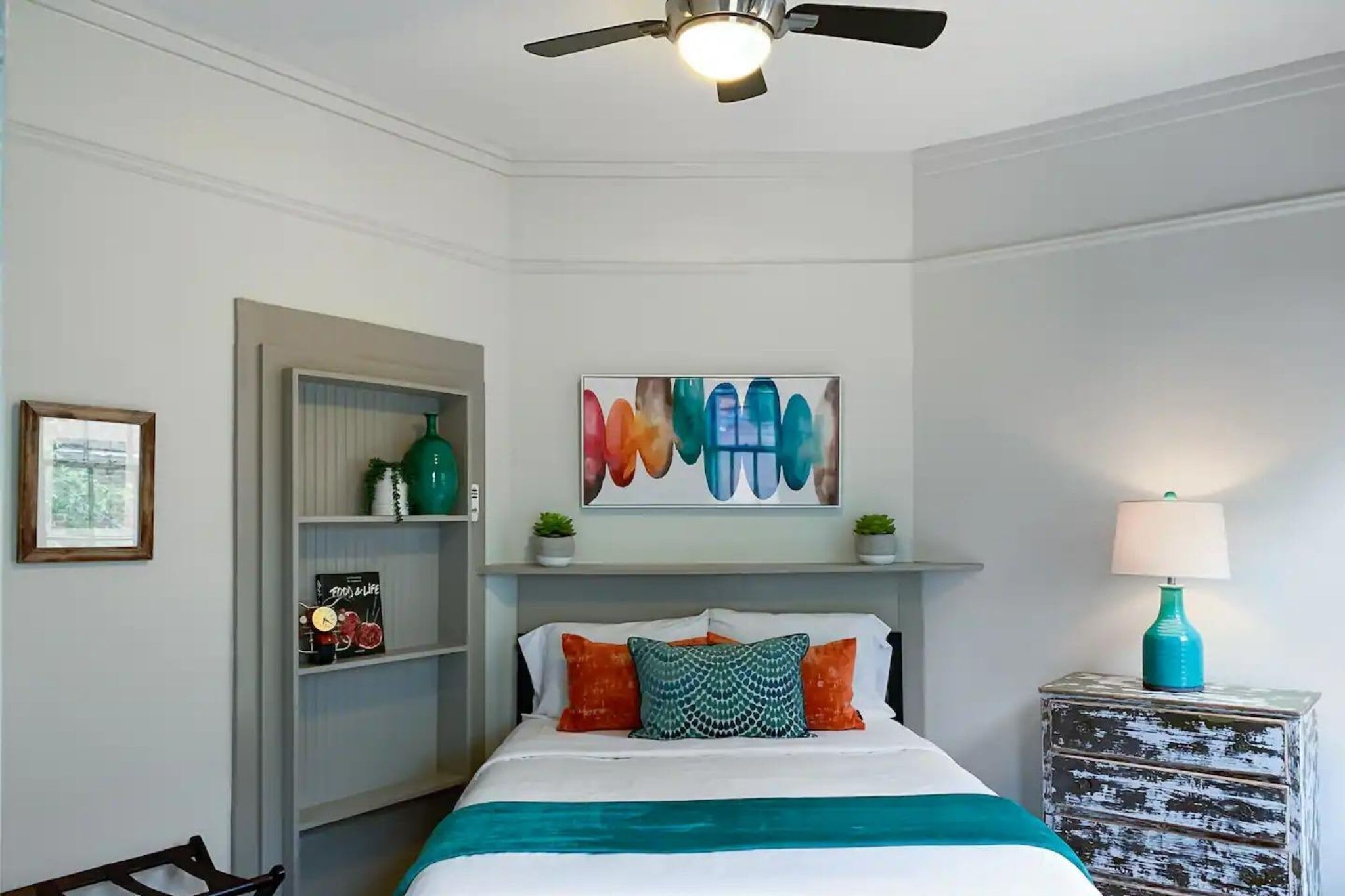A bedroom with a bed, a ceiling fan, a shelf with decorative items, and a wall-mounted artwork.
