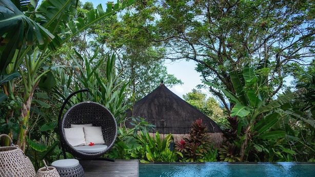 A wooden deck with a wicker chair and a small table, a pool, and lush tropical vegetation. - Palay (Malay)
