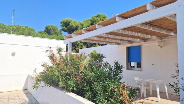 Un edificio blanco con una puerta azul y una pérgola de madera. Hay una silla blanca y una pequeña mesa cerca de la puerta.