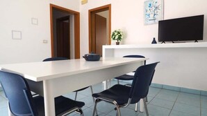 Un comedor con una mesa blanca, cuatro sillas, un televisor en un estante y una planta en maceta.