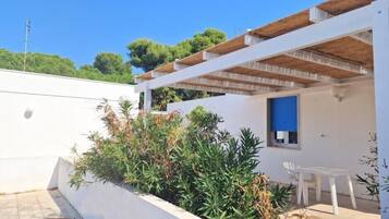 Un bâtiment blanc avec une porte bleue et une pergola en bois. On y trouve une chaise blanche et une petite table près de la porte.