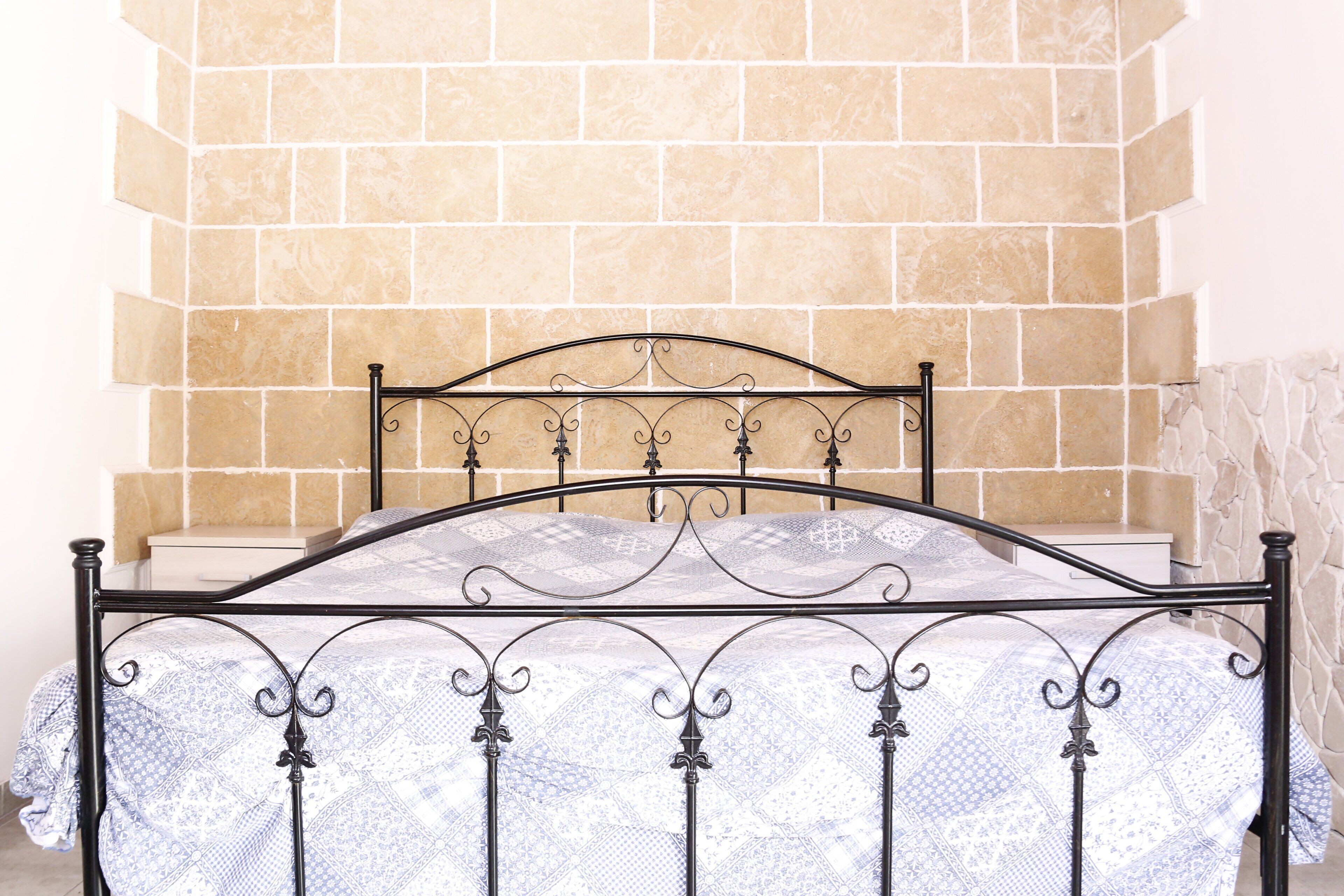 Una cama con estructura metálica y colcha estampada, pared revestida de piedra y una mesita de noche blanca con lámpara.