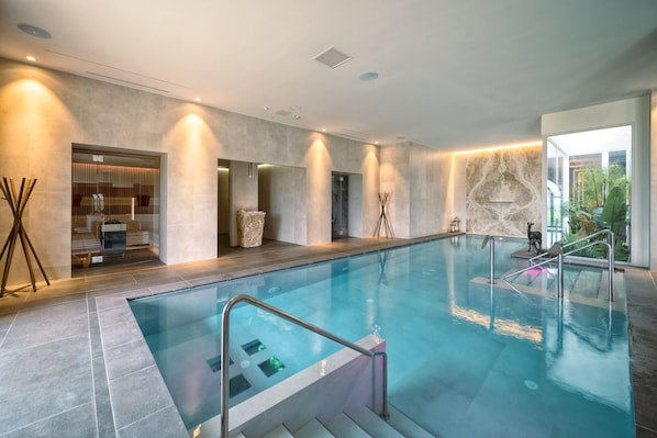 A modern indoor swimming pool with clear water, a glass barrier, and a stone wall. - The Club House (Castelnuovo Berardenga)