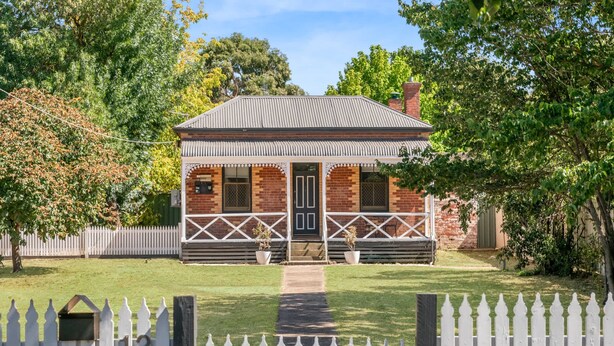 A small brick house with a porch, surrounded by a white picket fence and lush greenery.