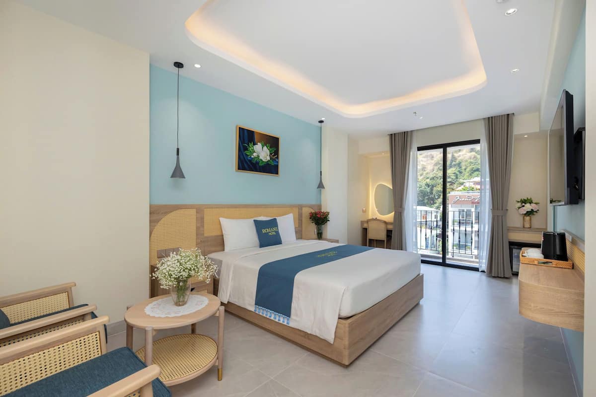 Una habitación de hotel moderna con una cama grande, una mesa redonda pequeña con un florero, una silla de mimbre y vistas al exterior a través de una puerta corrediza de vidrio.