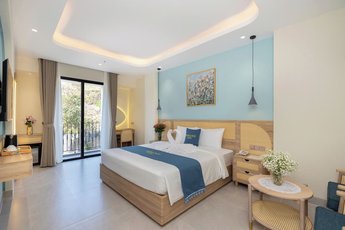 Habitación de hotel moderna con una cama grande, una mesita de noche, una pequeña mesa redonda con un florero y vistas al exterior a través de una puerta corredera.