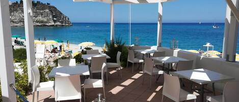 Un patio exterior con mesas y sillas blancas, con vistas a una playa y al mar.