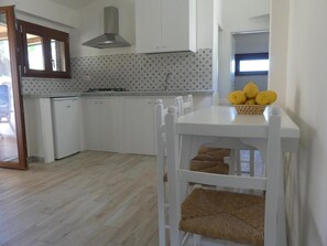 A modern kitchen with white cabinets, a gas stove, and a stainless steel range hood. There is a dining table with a woven chair and a basket of lemons on it. - Le Roccette Mare Beach Hotel & Restaurant (Tropea)
