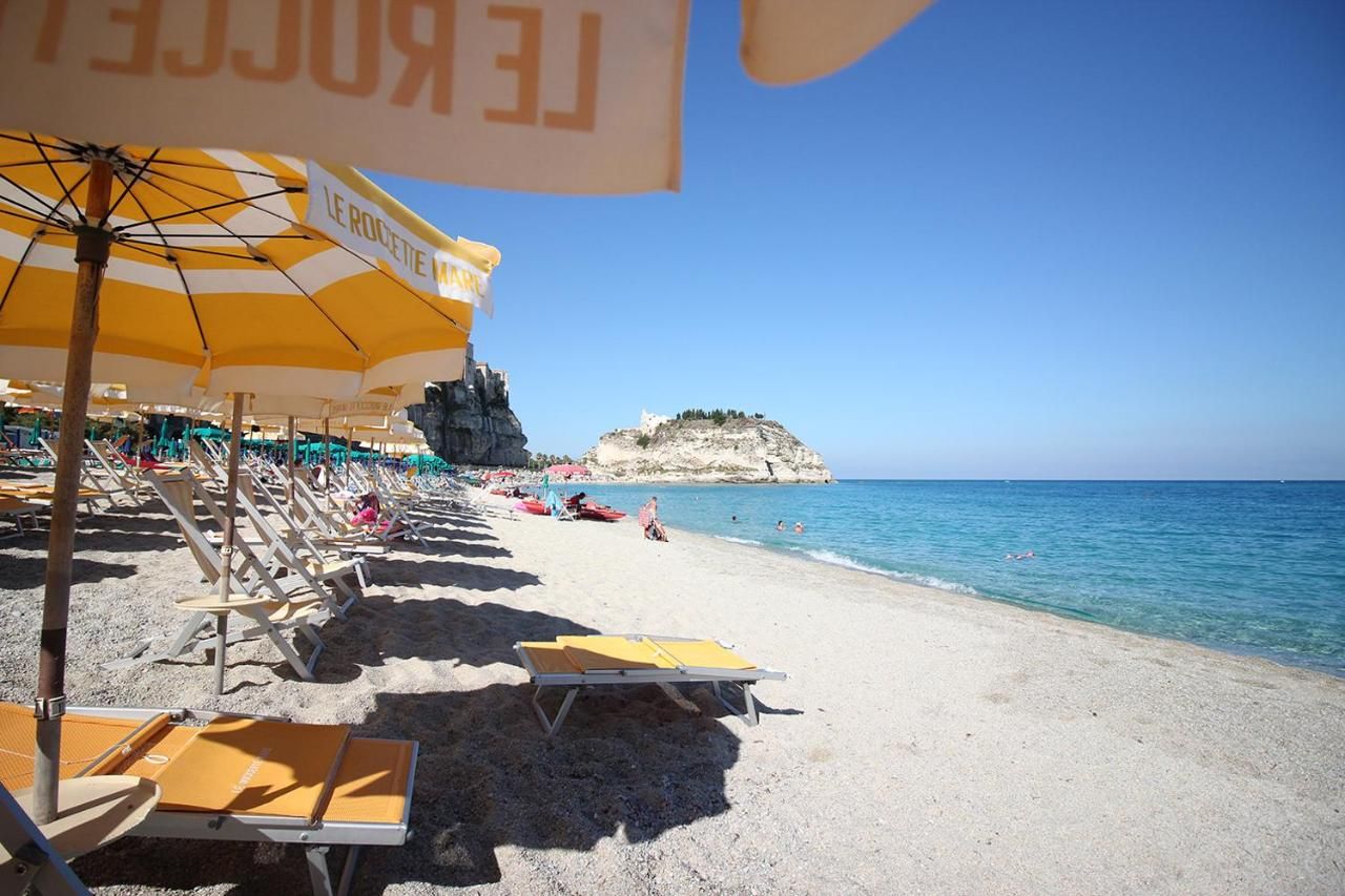 A beach with sun loungers and umbrellas, a clear sky, and a rocky outcrop in the background.