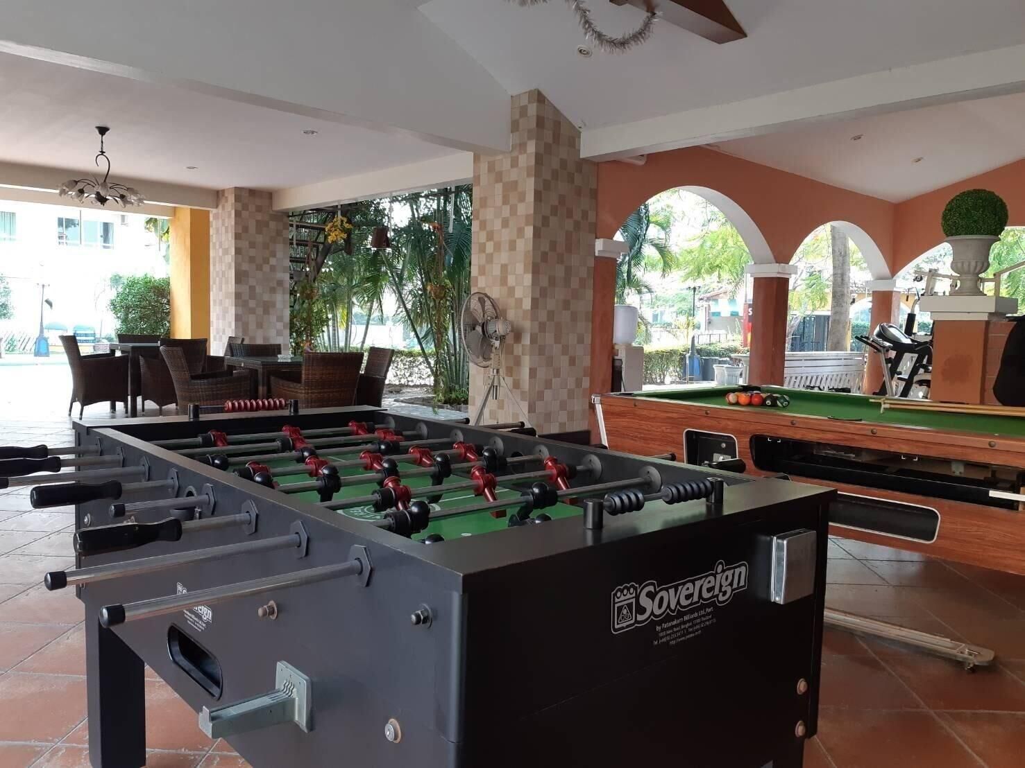 A foosball table labeled 'Sovereign' in a room with a pool table, seating area, and large windows overlooking a green outdoor area.