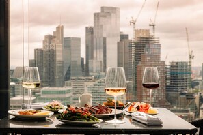 Ein Tisch mit verschiedenen Speisen und Weingläsern, vor einer Skyline einer Stadt.