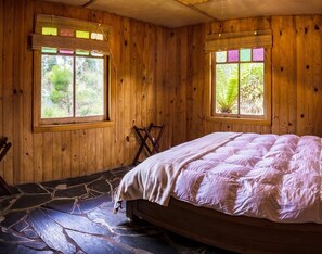 Une pièce en bois avec un lit, deux fenêtres équipées de stores et un sol en pierre.