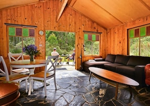L’intérieur d’un chalet en bois, avec un sol en pierre, une table à manger entourée de chaises, un canapé et de grandes fenêtres dotées de vitres teintées de vert.