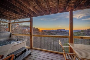 Une terrasse en bois avec un jacuzzi, une table et des chaises, offrant une vue panoramique sur un paysage magnifique au coucher du soleil.