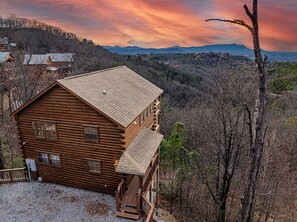 Une cabane en rondins à deux étages, dotée d’un balcon, entourée d’un paysage boisé et avec des montagnes en arrière-plan.