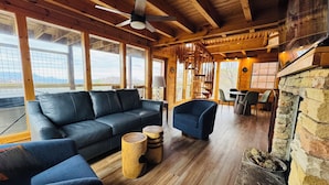 Un interno di una baita in legno con un divano, poltrone, un camino e ampie finestre.