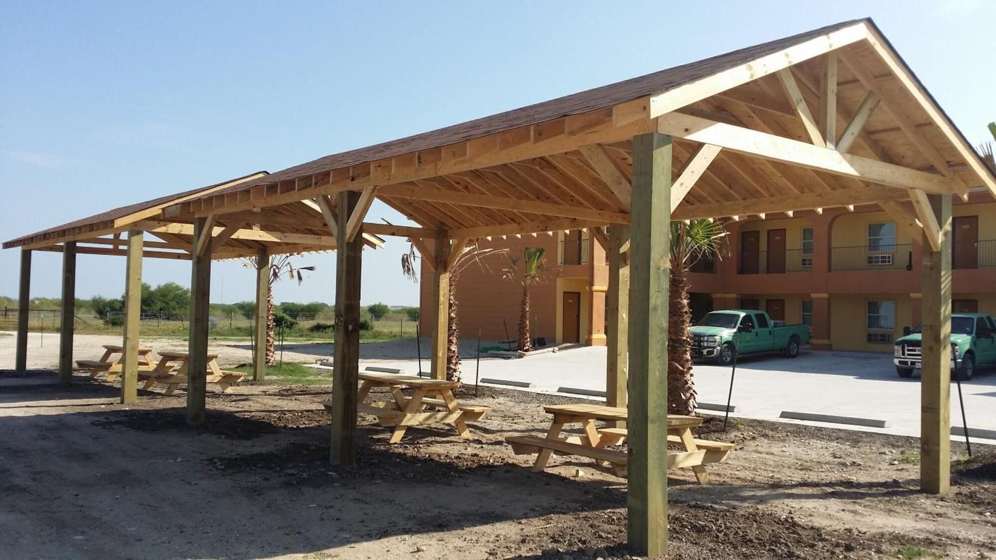 A covered outdoor seating area with picnic tables, a building in the background, and a clear sky.