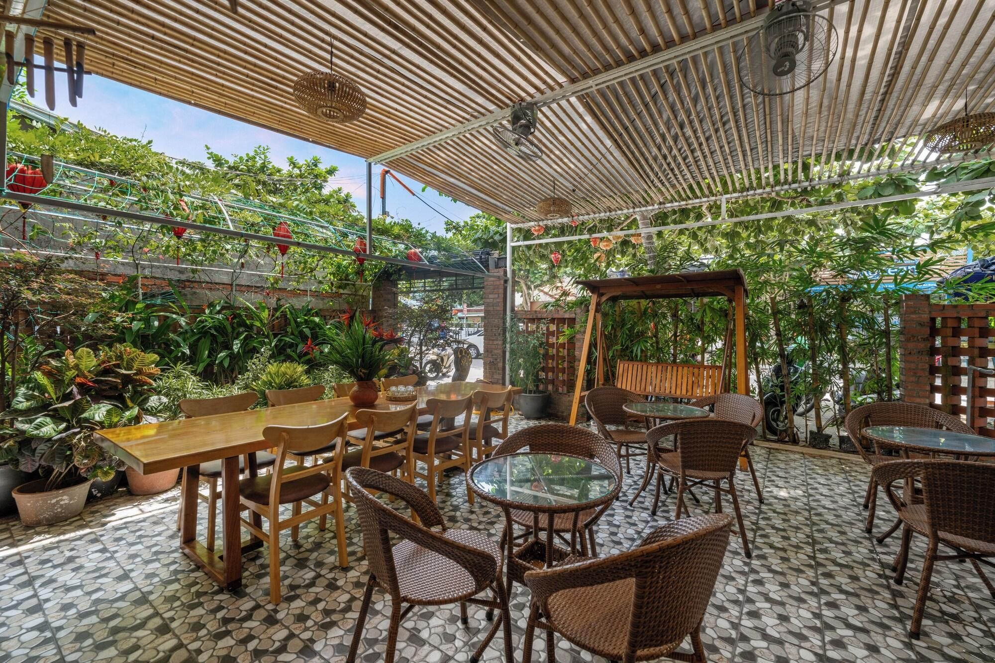 An outdoor cafe with wooden tables and wicker chairs, surrounded by potted plants and a bamboo ceiling.