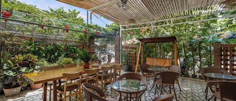 An outdoor cafe with wooden tables and wicker chairs, surrounded by potted plants and a bamboo ceiling.