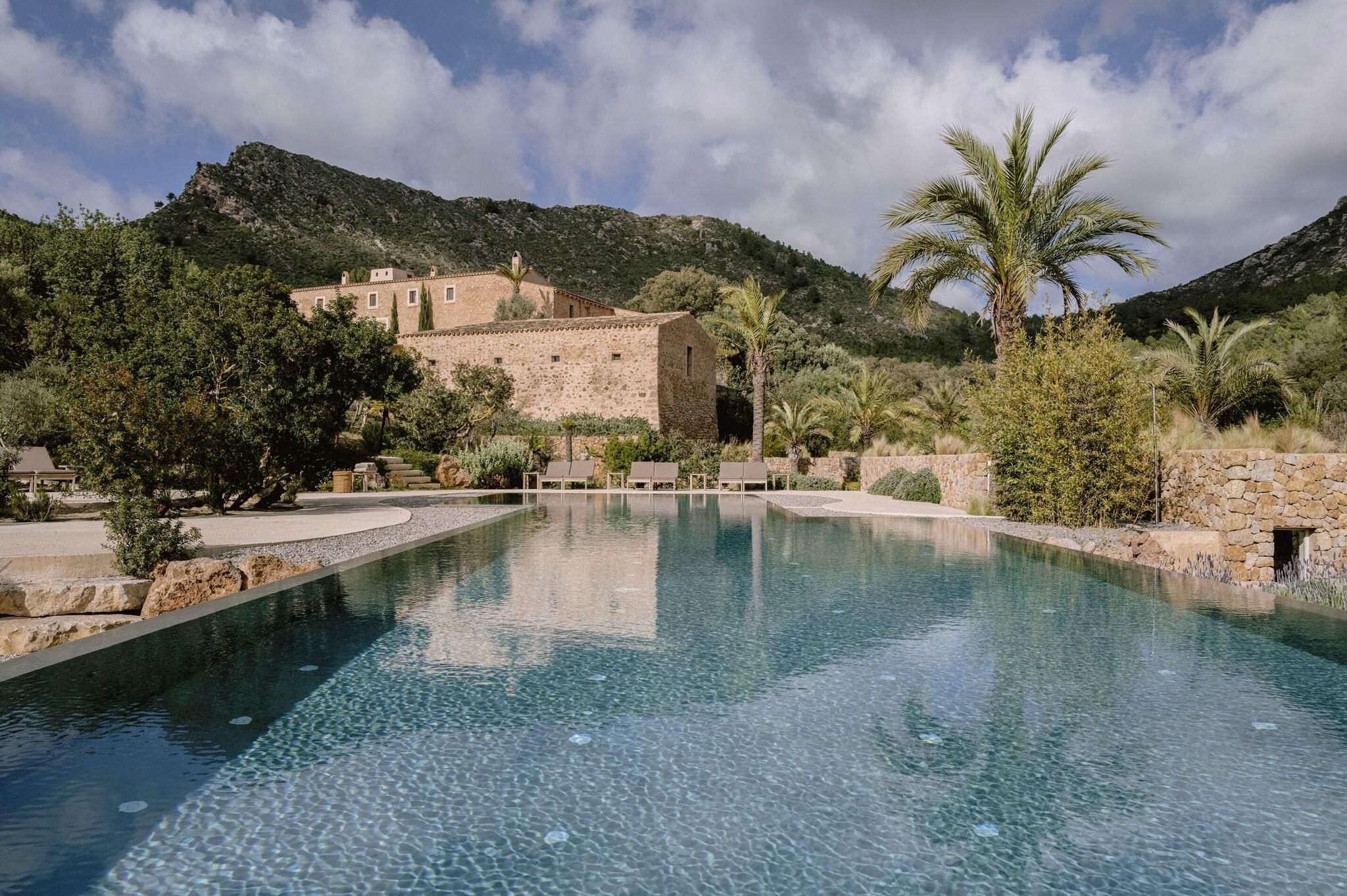 Una piscina cristalina rodeada de muros de piedra, con una montaña y un edificio al fondo.