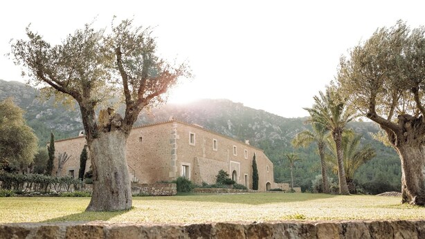 A rustic building with a stone facade, surrounded by trees and a stone wall, set against a mountainous backdrop. - Es Racó D Artá (Arta)