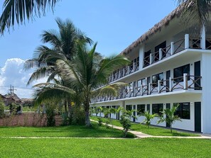 Un hotel moderno con facciata bianca, diversi balconi con ringhiere in legno e un prato curato con palme.
