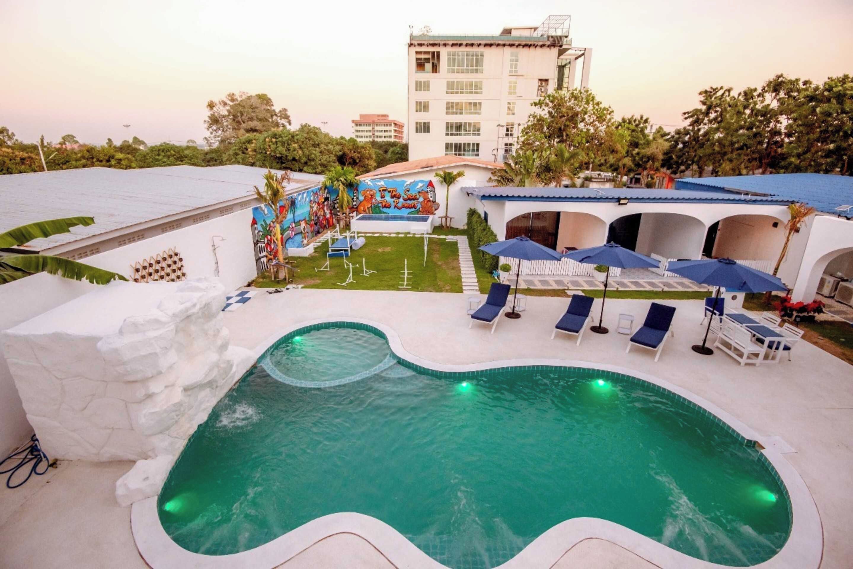 Une piscine entourée de chaises longues et de parasols, elle-même bordée d’un jardin aménagé et d’un bâtiment orné d’une fresque murale.