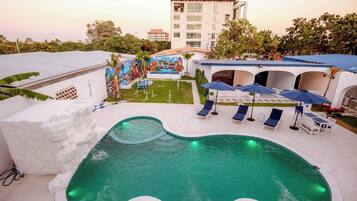 A swimming pool with lounge chairs and umbrellas, surrounded by a landscaped garden and a building with a mural.