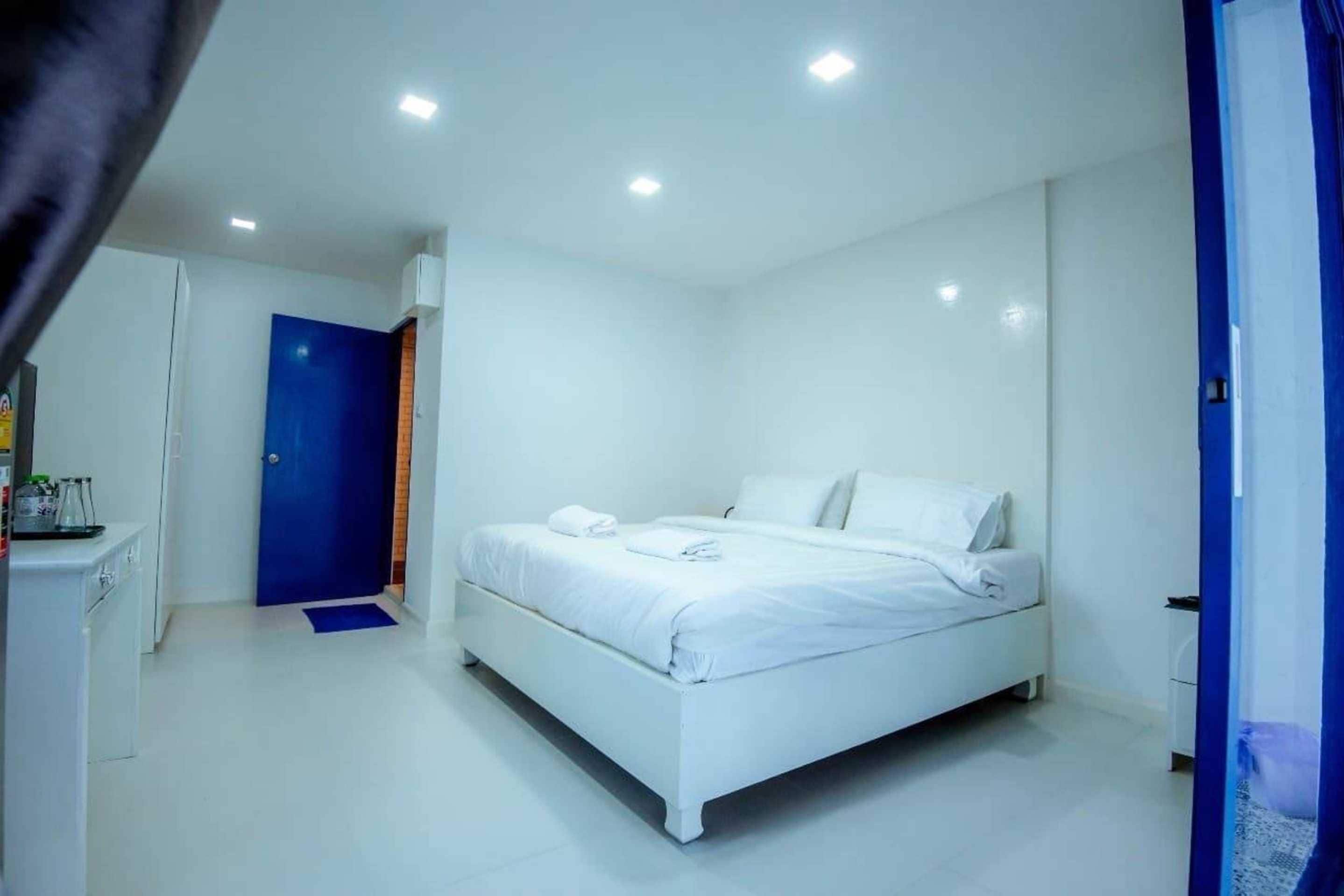 A modern, white-themed bedroom with a single bed, a blue door, and a small desk with a chair.