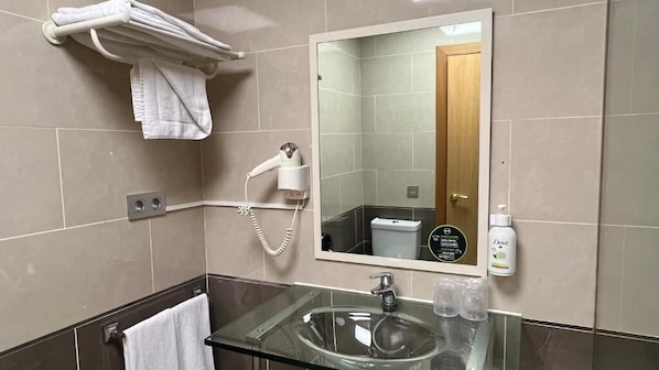 Un baño moderno con un lavamanos de vidrio, un secador de pelo, un perchero para toallas y una botella de Dove.
