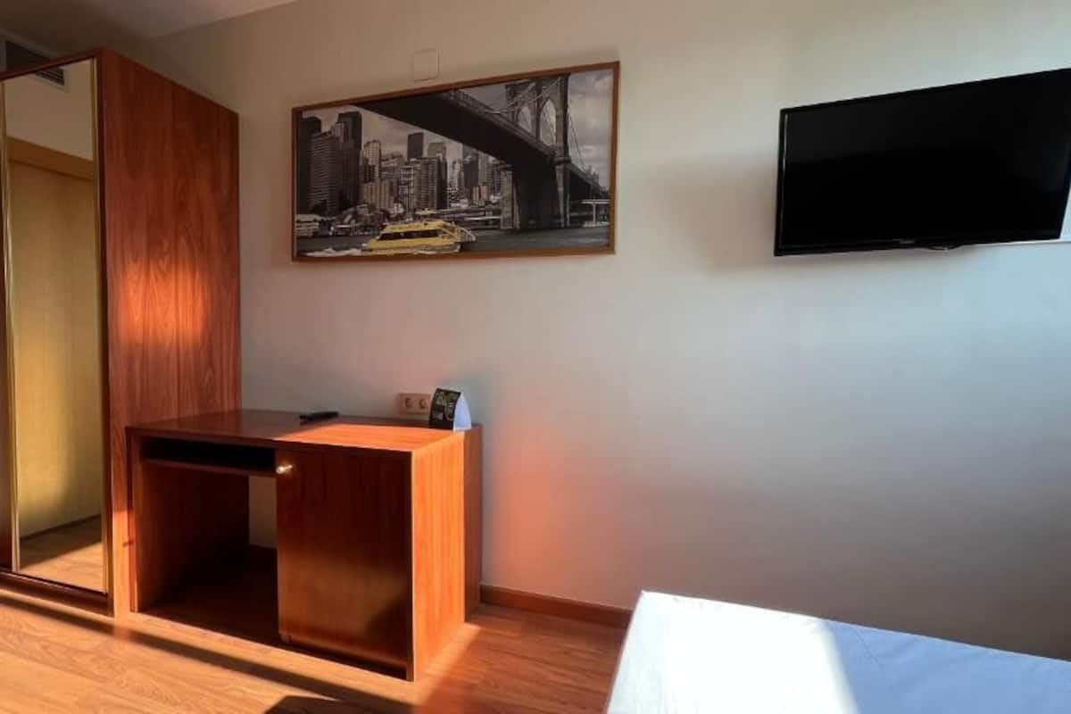 A room with a wooden desk, a mirror, a framed cityscape picture, and a TV on the wall.